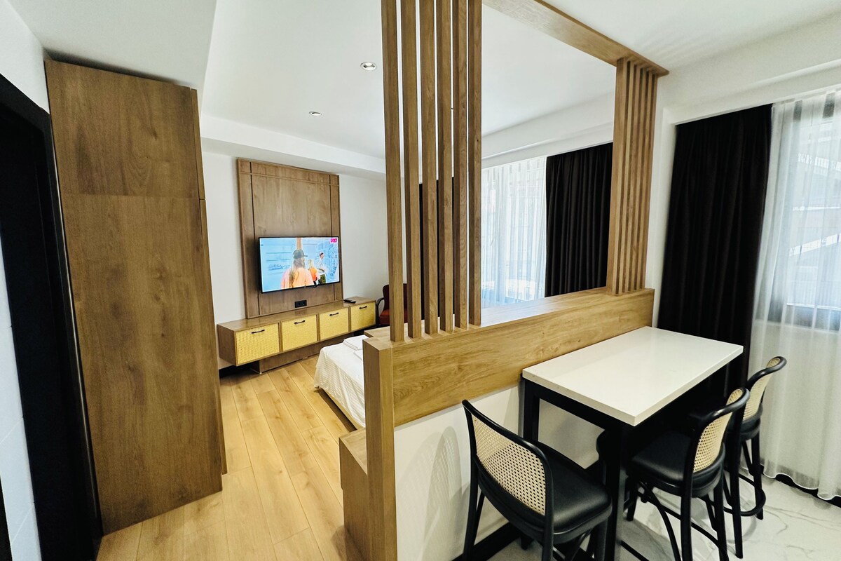 A modern hotel room with a wooden partition, a TV on the wall, a bed, and a dining area with chairs.