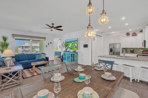 A modern living room with a blue sofa, wooden coffee table, and a dining area with a wooden table set for four. There is a kitchen with white cabinets and a large island.