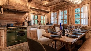 Un intérieur rustique en bois, avec une table à manger dressée pour le repas, un poêle d’époque et de grandes fenêtres.