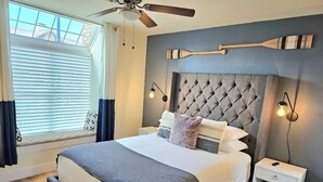 Ein Schlafzimmer mit einem gesteppten Kopfteil, einem Bett mit grau-weißer Bettwäsche, einem Nachttisch mit Lampe und einem Fenster mit Jalousien.