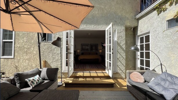 An open doorway leading to a bedroom with a bed, two bedside lamps, and framed pictures on the wall. There is a large umbrella providing shade over a seating area with a sofa and a chair. - Santa Monica 3 bedroom house with sunny outdoor deck (Los Angeles)