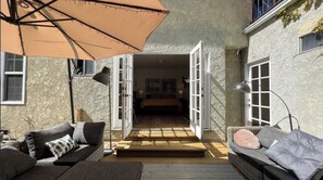 Un umbral abierto da paso a un dormitorio con cama, dos lámparas de noche y cuadros enmarcados en la pared. Un paraguas grande proporciona sombra a una zona de estar con sofá y sillón.