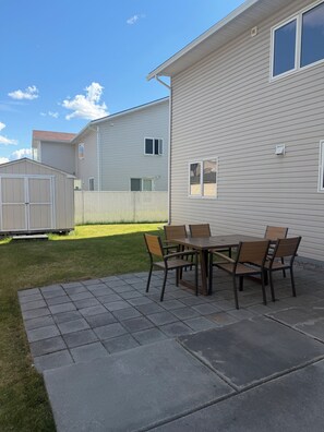 Ein Wohngrundstück mit einer Terrasse, einem Tisch und Stühlen, einem Gartenhaus und einem Haus mit Verkleidung und Fenstern.