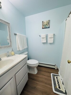 Ein Badezimmer mit einem weißen Waschbecken, einer Toilette und einer Duschvorhang. Handtücher hängen an der Wand, und über der Toilette hängt ein kleines Bild.
