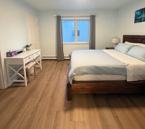 Ein Schlafzimmer mit einem hölzernen Bett, einem Schreibtisch und einem Stuhl. Ein Fenster mit Vorhängen bietet Blick ins Freie.