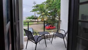 A balcony with two chairs, a red car, and a view of a green area with trees and a power line. - Pousada Nascimentos (Bertioga)