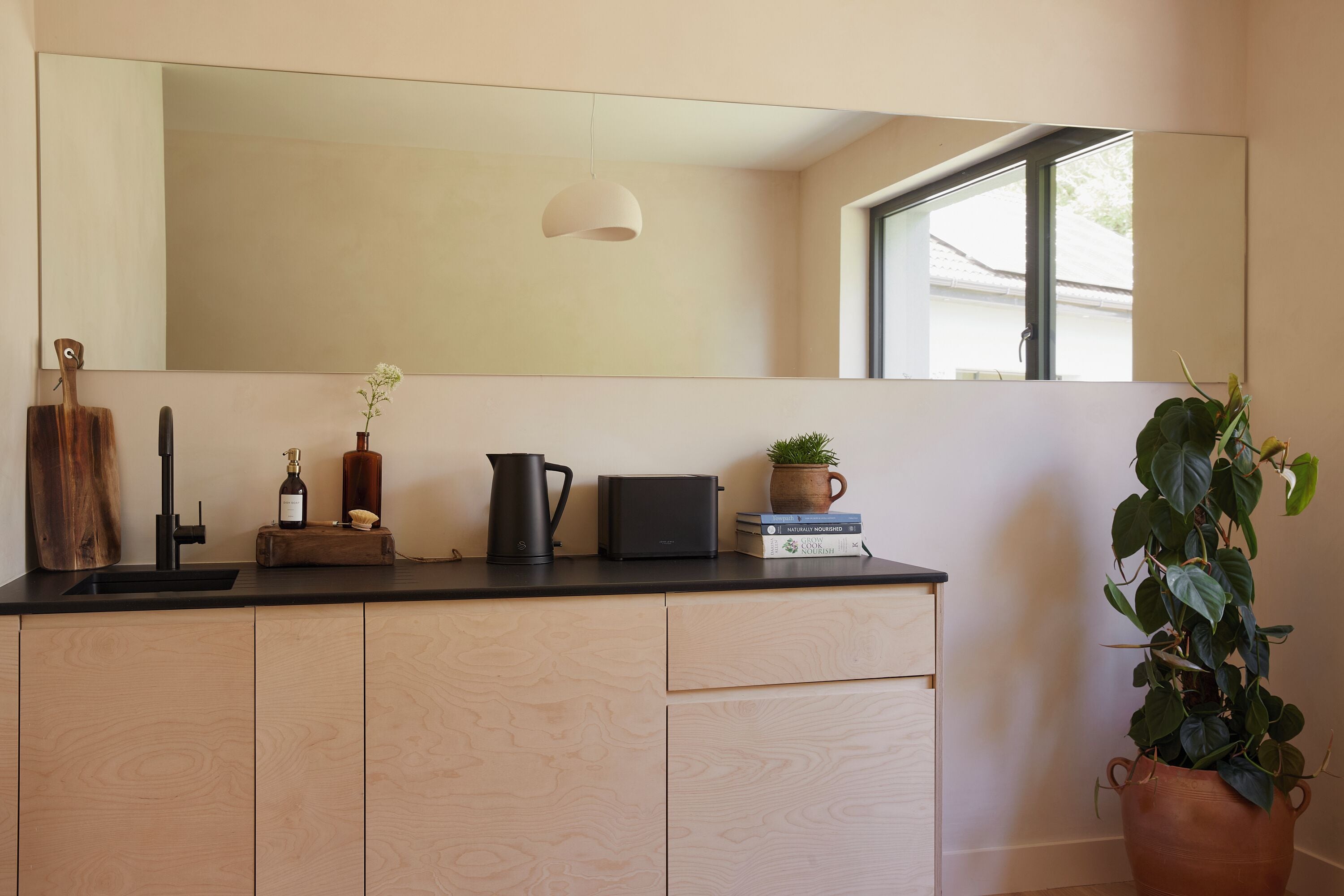 Een keuken met een zwart aanrecht, houten kastjes, een grote spiegel, een hanglamp en een potplant.