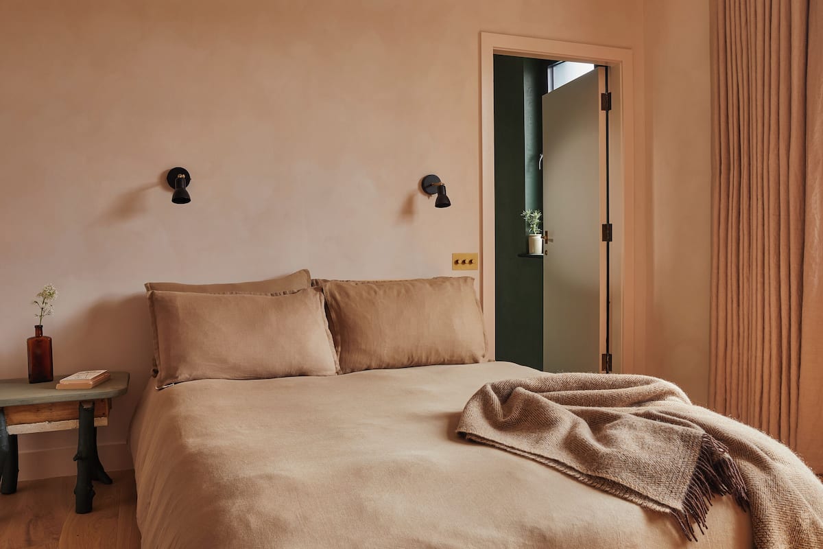 A bedroom with a bed, two wall-mounted light fixtures, a bedside table with a book and a vase, and a door leading to another room.