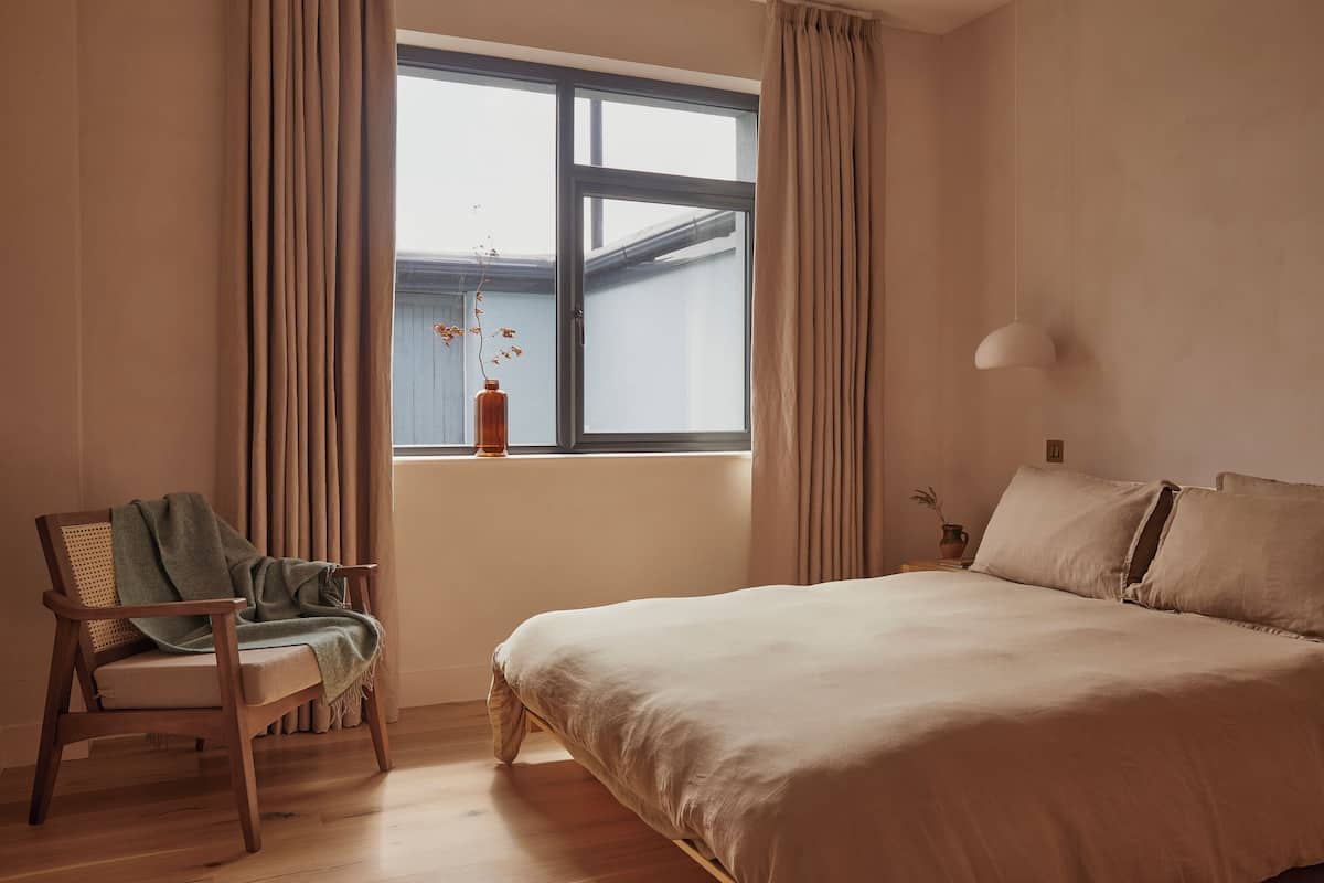 A bedroom with a large bed, a wooden chair with a blanket, a window with curtains, and a vase with plants.