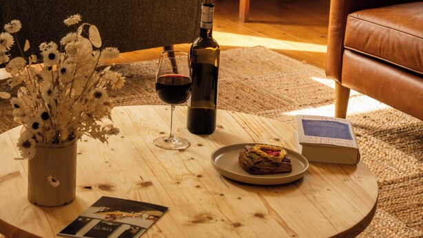 A cozy room with a bed, a wooden coffee table with a bottle, wine glass, and a book, and a leather chair.