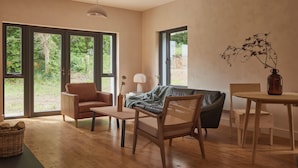 A modern living room with a leather sofa, wooden chairs, a small table, and large windows offering a view of greenery.
