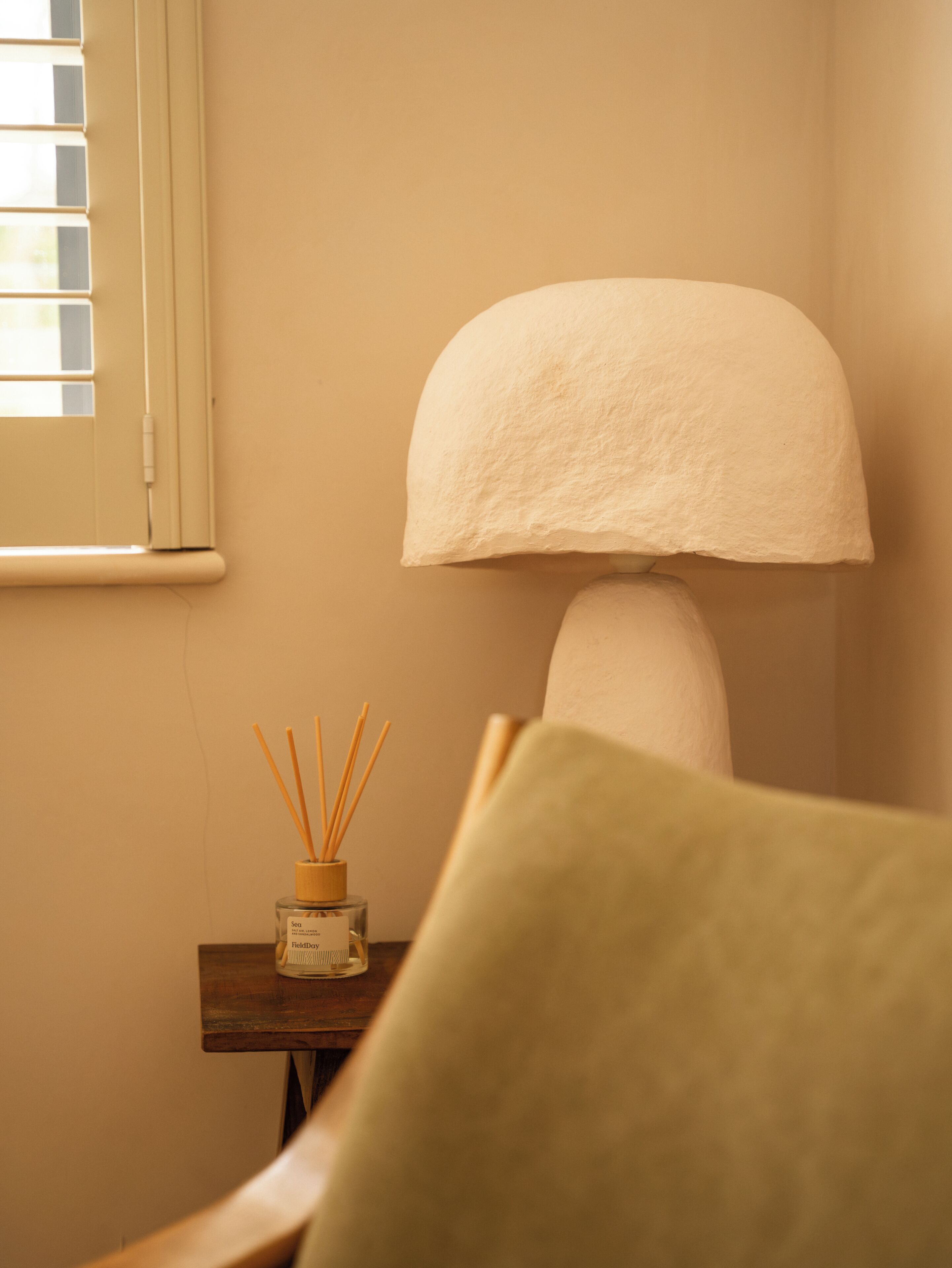 A room with a lamp, a small table with a bottle and reed sticks, and a chair.
