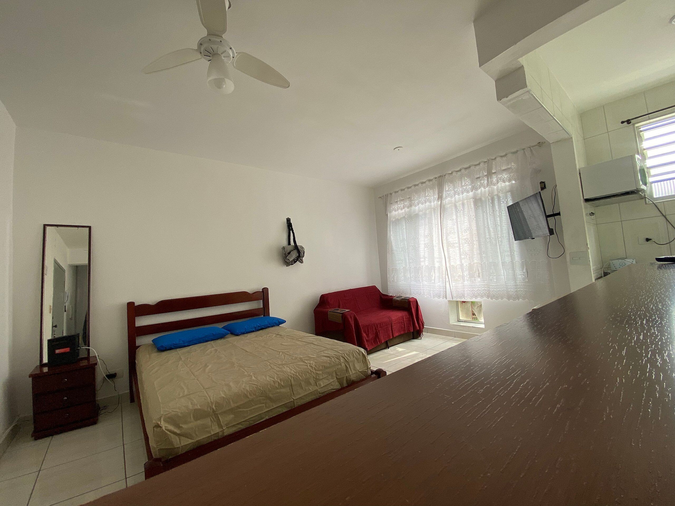 Un dormitorio con cama, una silla roja, ventilador de techo, ventana con cortinas y una pequeña mesa.