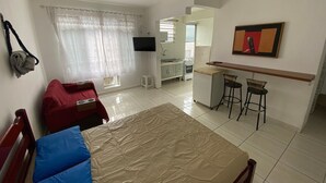 A bedroom with a bed, a desk with two chairs, a TV, and a kitchenette. - Pé na Areia Canto do Forte (Praia Grande)