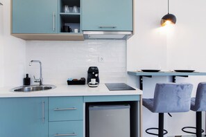 A modern kitchen with teal cabinets, a stainless steel sink, a coffee maker, and a dishwasher. There are two bar stools and a pendant light above the counter.