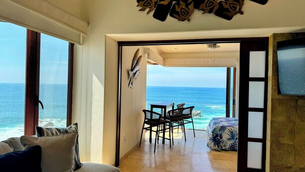 A room with a view of the ocean, a dining area with chairs and a table, and a bed with a floral bedspread. - NEW LISTING! OCEANFRONT CORNER TOP FLOOR 2 BEDROOM UNFORGETTABLE VIEWS
 (San Lucas)
