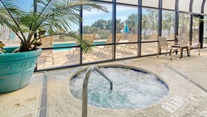 An indoor pool area with a hot tub, lounge chairs, and a large window offering a view of the pool and palm trees.