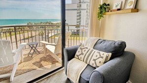 A balcony with a view of the beach, a grey sofa with patterned cushions, and wooden shelves with decorative items.