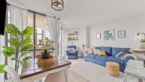 A modern living room with a wooden dining table, blue sofa, and large windows with white curtains.