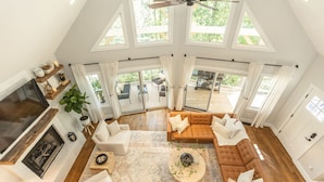 A spacious living room with a fireplace, a large sectional sofa, and a coffee table. There are multiple windows providing natural light and a view of the outdoors. - Cozy Luxury Treehouse, 10 Min To Asheville, Mountain Views (Swannanoa)