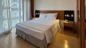 Une chambre d’hôtel moderne avec un grand lit, une table de chevet avec une lampe, une petite table de nuit avec un téléphone et une vue sur la ville grâce à de grandes fenêtres.