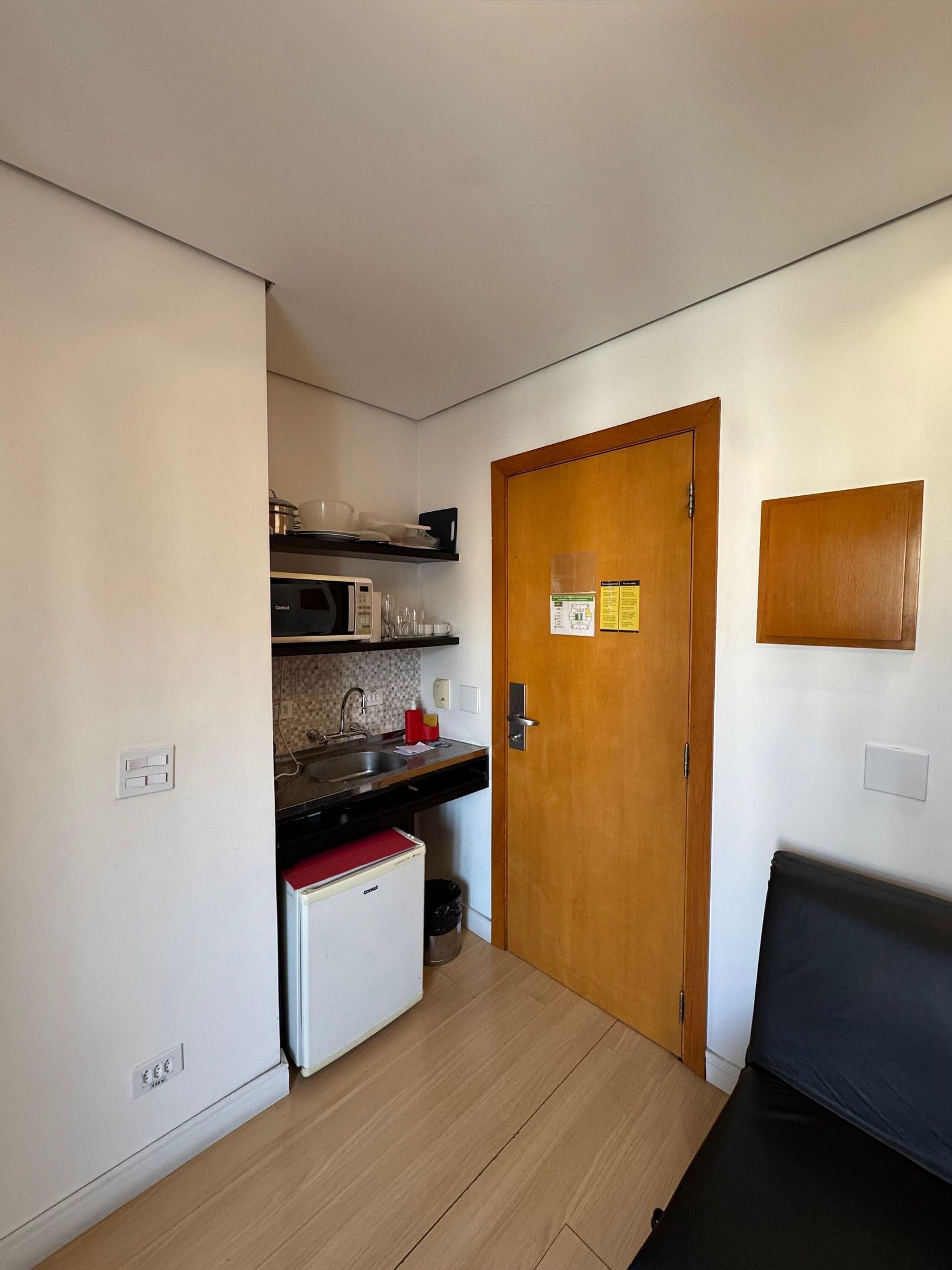 A compact kitchen with a microwave, sink, and small refrigerator. A wooden door with a notice is visible, and a bench is placed in the room.