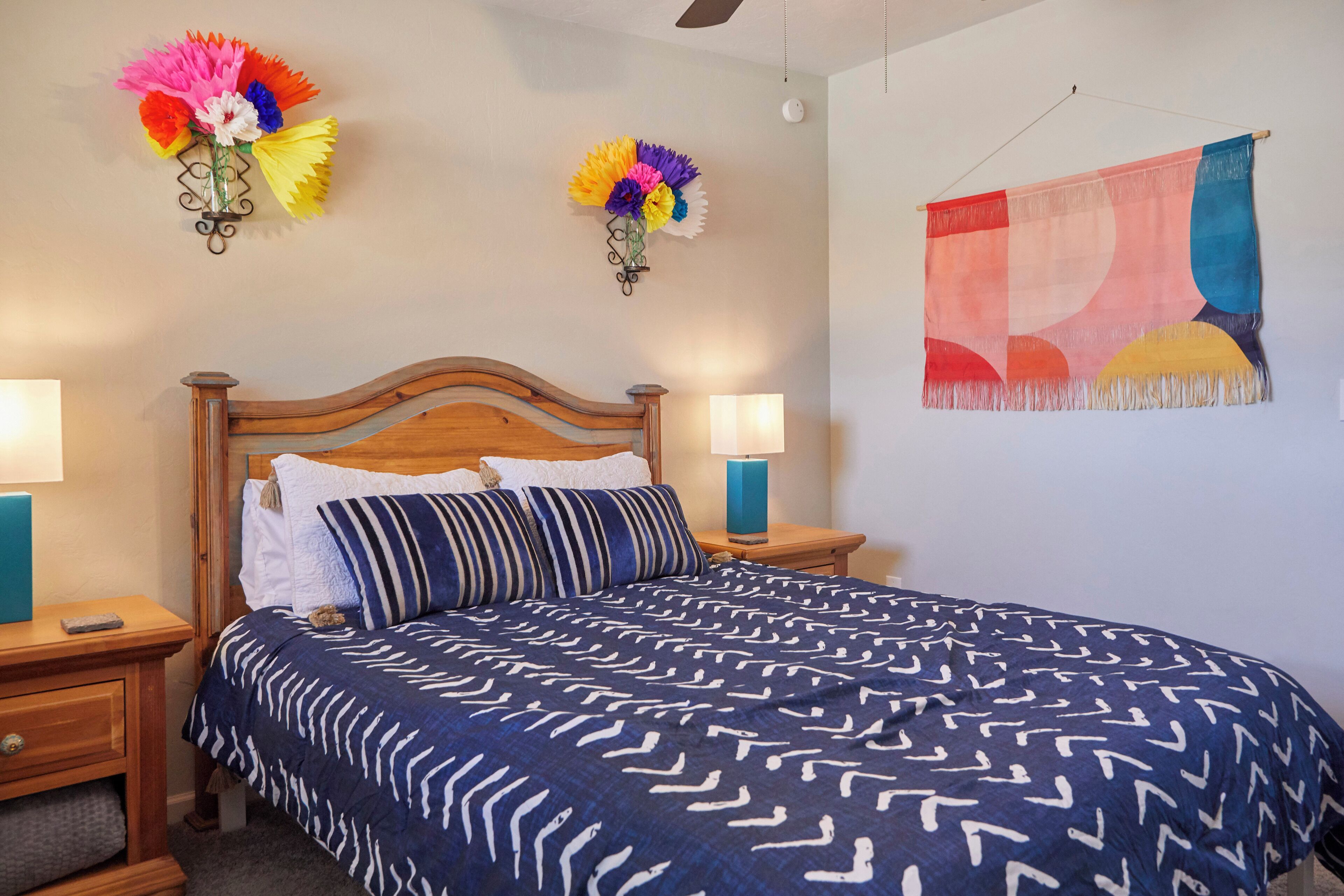 A bedroom with a wooden headboard, two bedside tables with lamps, a bed with a patterned blue blanket, and a colorful abstract painting on the wall.