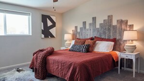 A bedroom with a large bed, two bedside tables with lamps, a cityscape wall art, and a window.