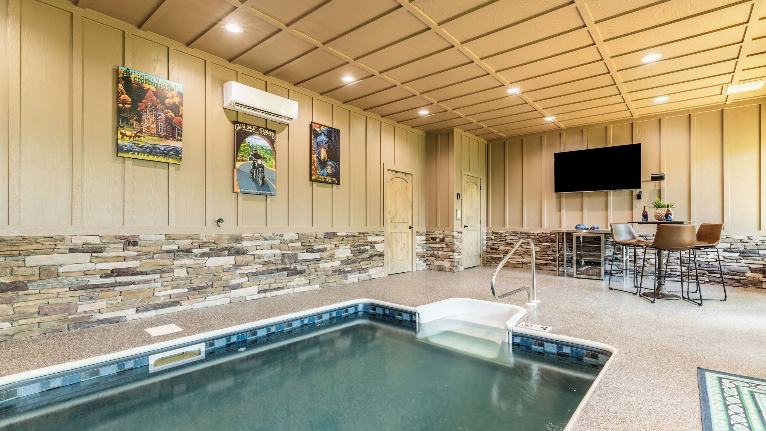 An indoor pool area with stone walls, a mounted TV, and a bar area with chairs.