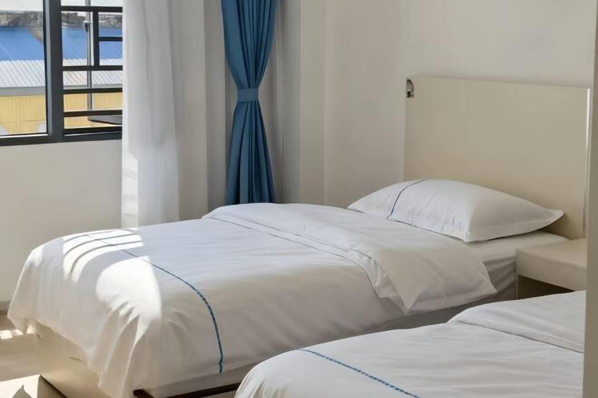 A hotel room with two beds, a window with curtains, and a view of a blue building outside.