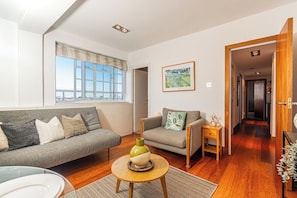A modern living room with a grey sofa, a smaller grey armchair, a round coffee table with a vase, and a wooden side table. A large window with a view of the city is visible, and a framed picture hangs on the wall.