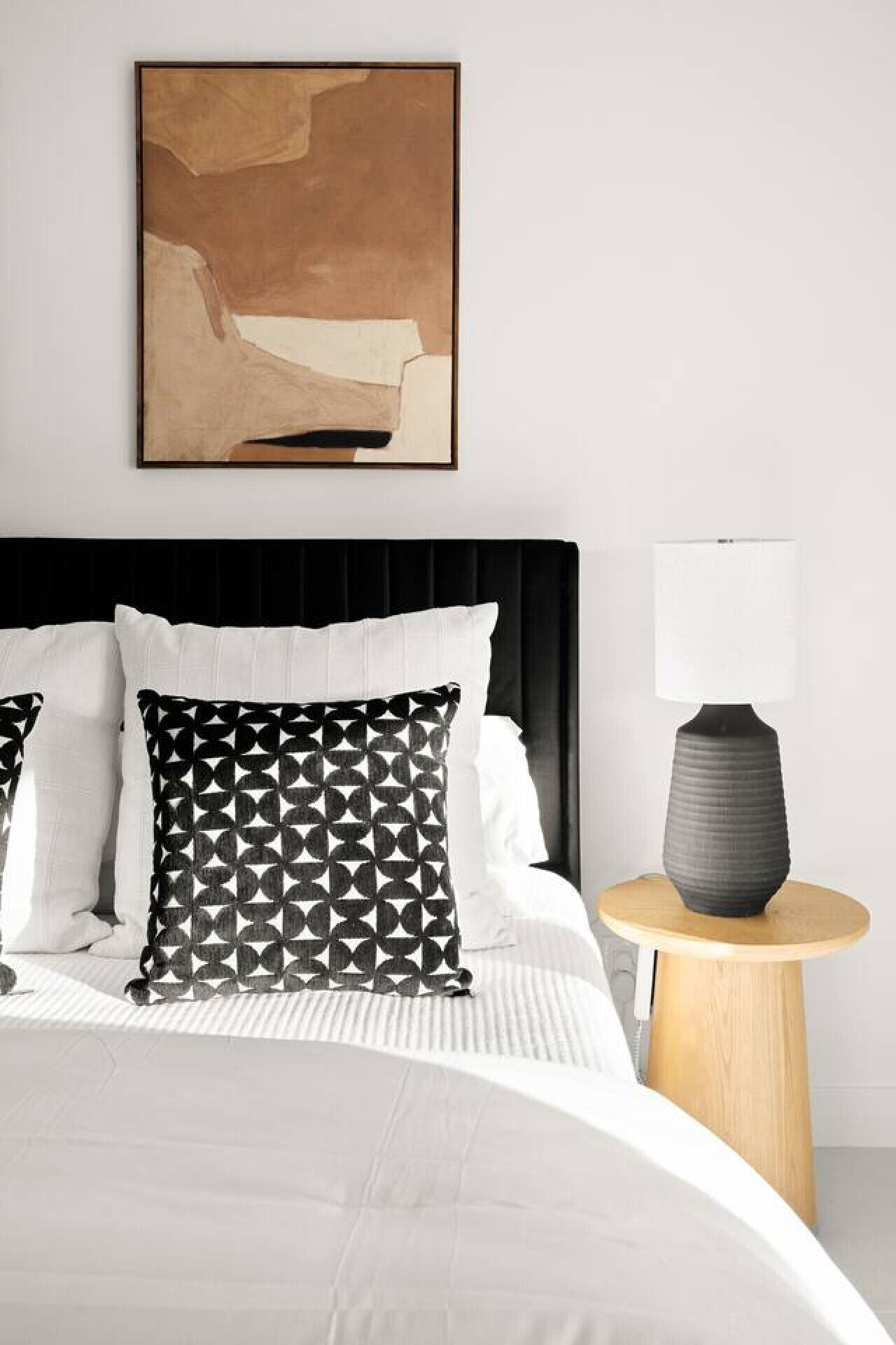 Een moderne slaapkamer met een groot bed, zwart-wit gestreepte kussens, een zwart hoofdbord, een witte nachtkastje met een lamp, en een minimalistisch abstract schilderij aan de muur.