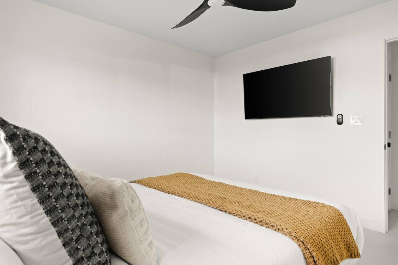 Ein modernes Schlafzimmer mit einem Einzelbett, einem Deckenventilator, einem an der Wand montierten Fernseher und einer Tür mit Gegensprechanlage.