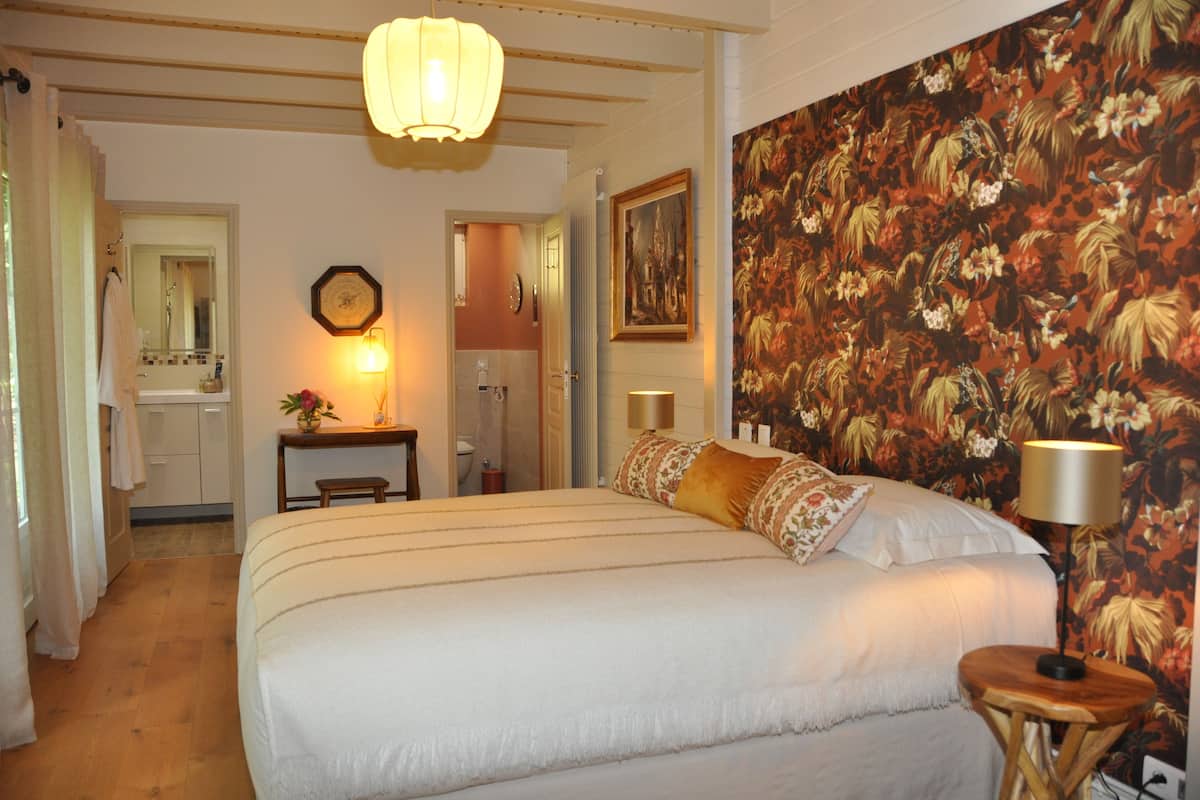 Une chambre avec un grand lit, une table de chevet, une lampe, un miroir, une porte donnant sur une salle de bain et un papier peint à motifs floraux.