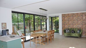 A spacious dining area with a wooden table and chairs, large windows offering a view of greenery, and a green sofa with floral upholstery in the background.