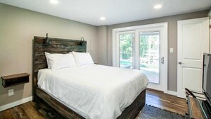 A bedroom with a wooden headboard, a large bed with white bedding, a small bedside table, and a sliding glass door leading to an outdoor area.