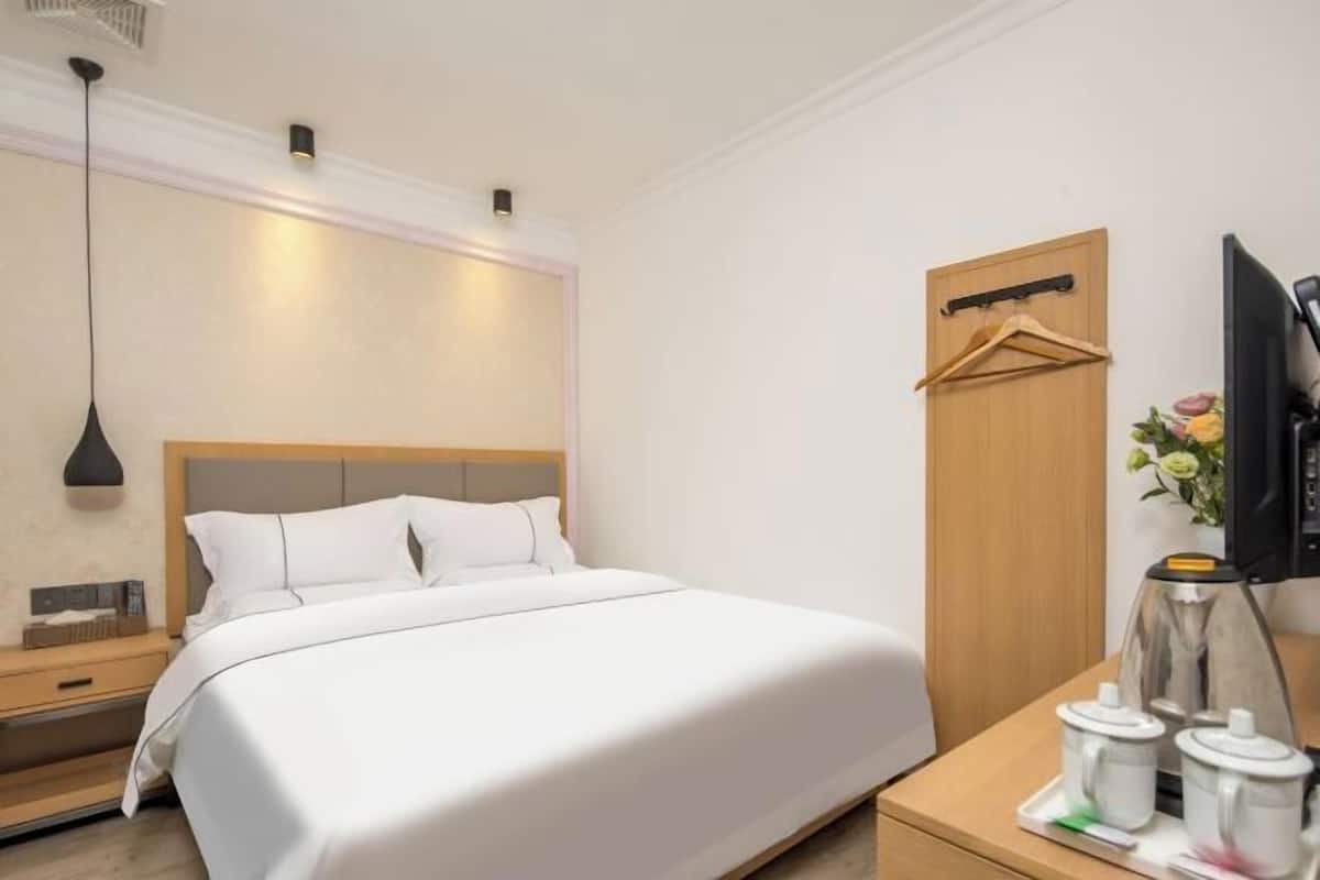 Quarto de hotel com cama, mesa de cabeceira, televisão e cabideiro fixado na parede.