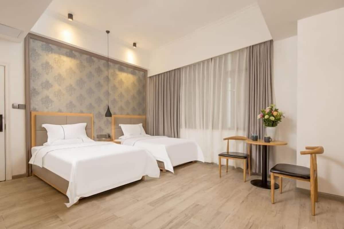 Quarto de hotel com duas camas, uma escrivaninha, uma cadeira, uma luminária e uma janela grande com cortinas.