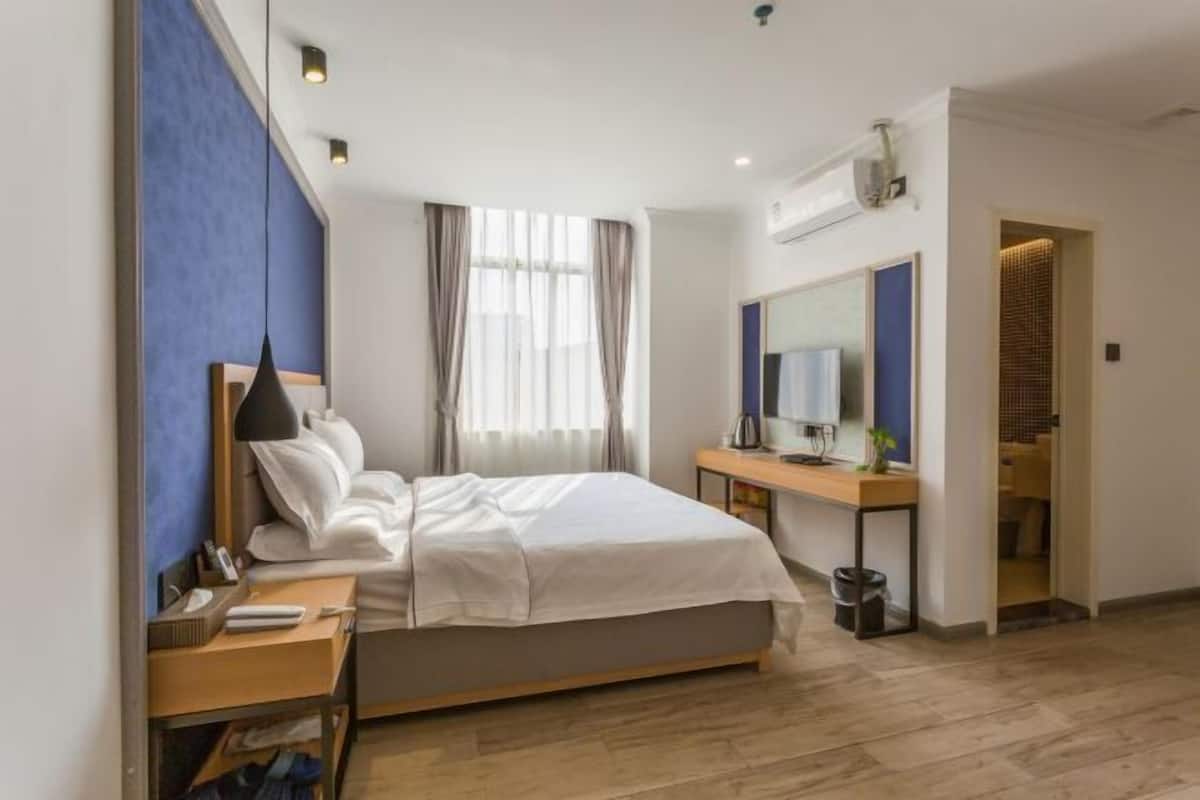Quarto de hotel moderno com cama, mesa de cabeceira e secretária com cadeira. Há uma janela com cortinas, um espelho e um ar condicionado fixado na parede.