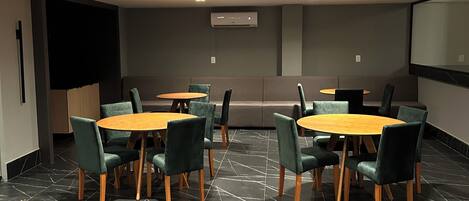 A conference room with two round tables, green upholstered chairs, a projector screen, and a wall-mounted air conditioning unit.