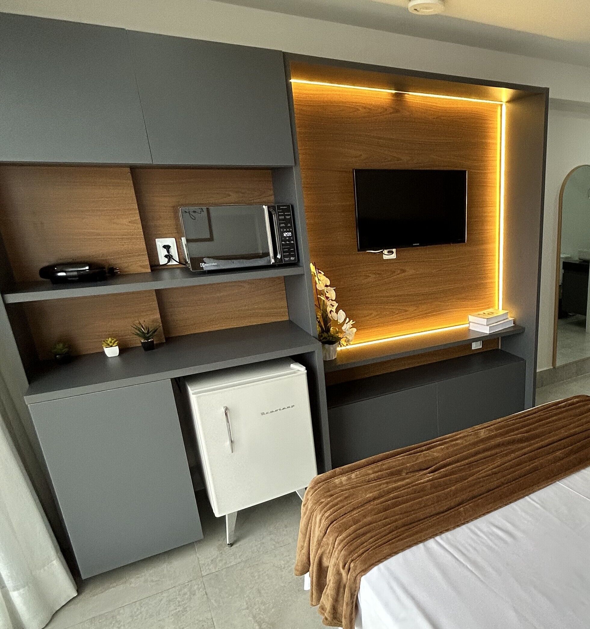 A modern bedroom with a built-in desk, a microwave, a flat-screen TV, and a bedside table with a lamp.