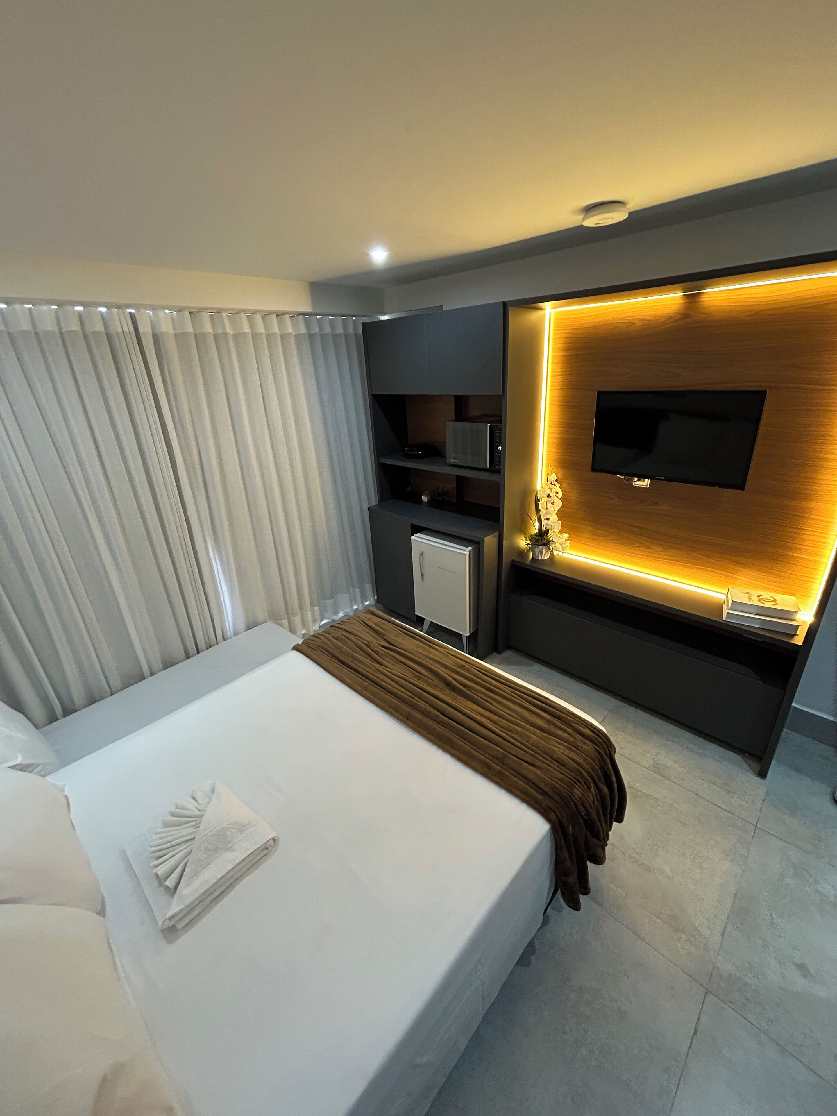 A modern hotel room with a large bed, a flat-screen TV mounted on a wooden wall, a small desk with a chair, and a curtain-covered window.