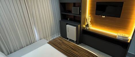 A modern hotel room with a large bed, a flat-screen TV mounted on a wooden wall, a small desk with a chair, and a curtain-covered window.