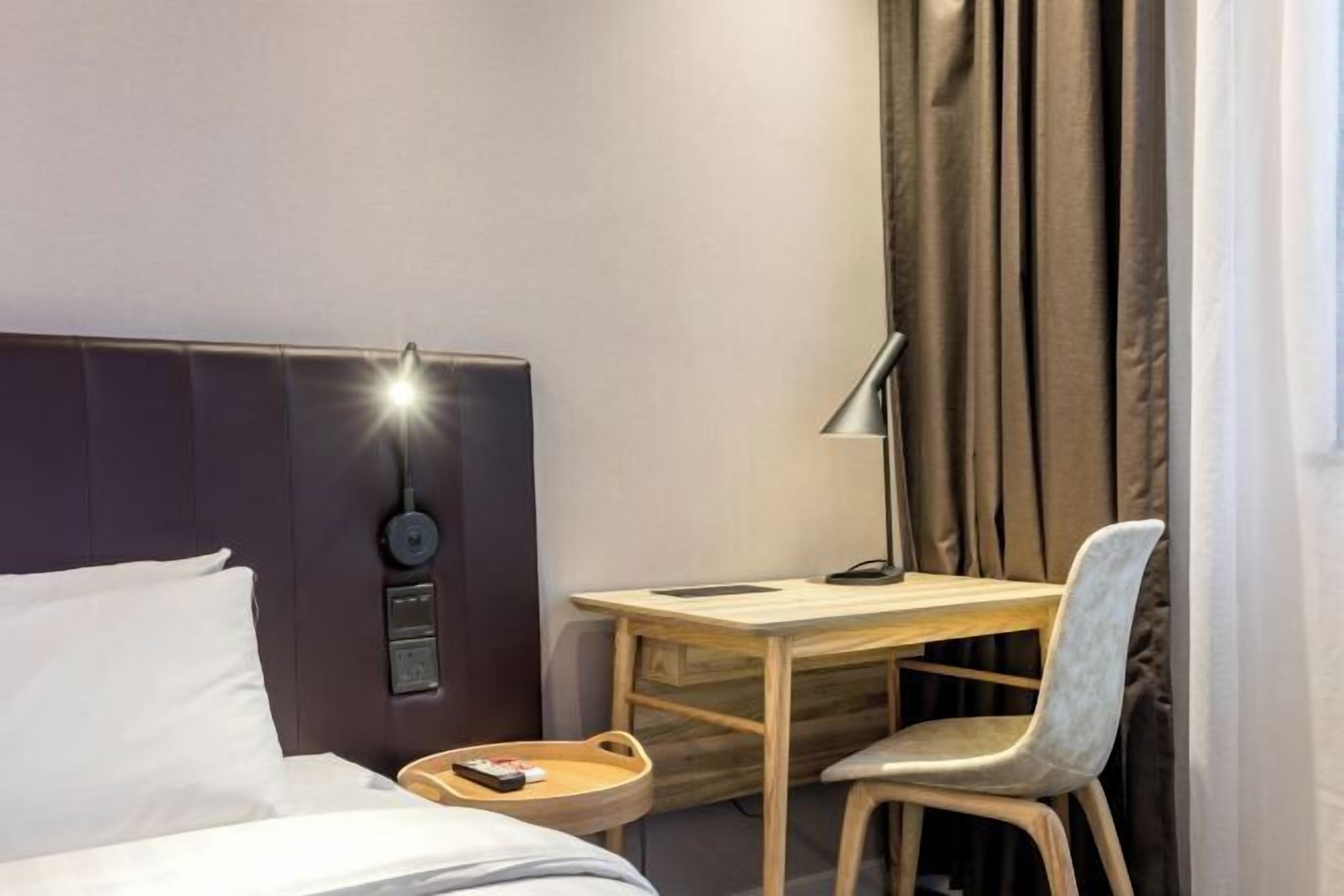 Hotellihuone, jossa on sänky, työpöytä, tuoli, lamppu, yöpöytä ja verhot.