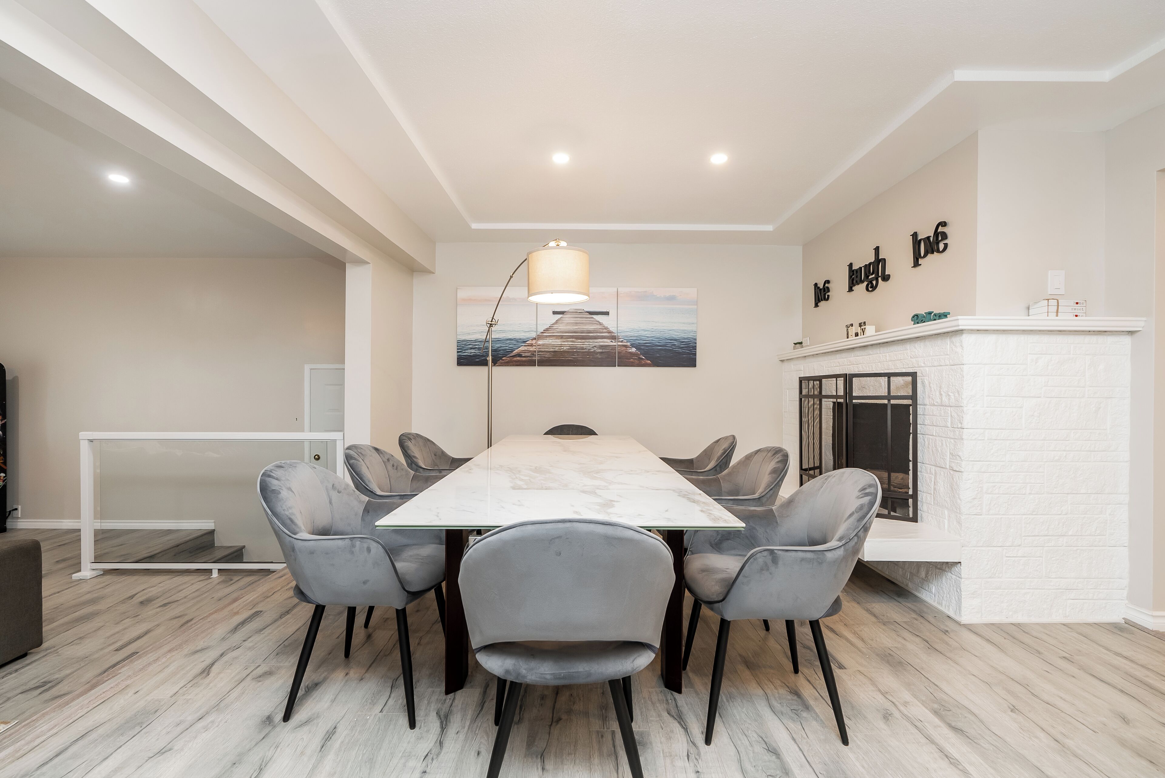 A modern dining area with a marble table, grey upholstered chairs, a white brick fireplace, and a painting of a pier on the wall.