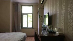 A hotel room with a bed, a desk with a computer, a window with curtains, and a wall-mounted air conditioner.