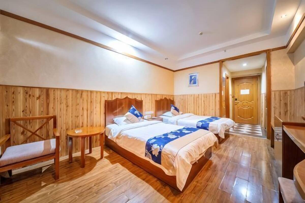 A hotel room with two beds, wooden flooring, a wooden headboard, a small table, and a bench.