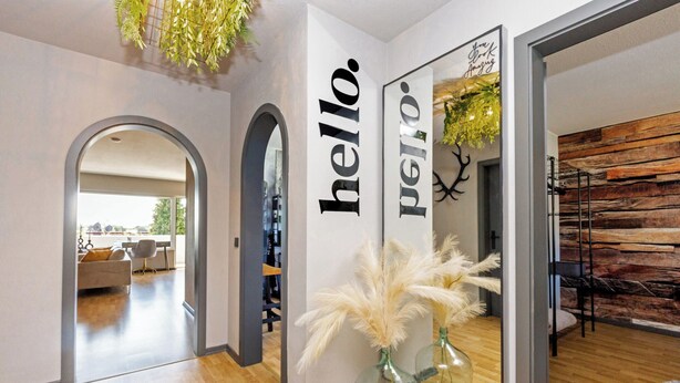 A modern interior with a hallway leading to a living area, featuring a mirror with 'hello' and 'polo' text, a glass vase with white feathers, and a wooden wall. - Modern apartment with balcony & 2 parking spaces (Odenthal)