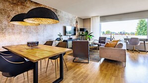 A modern living room with a wooden dining table, comfortable seating, a large window with a city view, and a TV mounted on the wall. - Modern apartment with balcony & 2 parking spaces (Odenthal)
