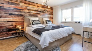 A modern bedroom with a wooden wall panel, a large bed with pillows, a bedside table with a lamp, and a window with curtains. - Modern apartment with balcony & 2 parking spaces (Odenthal)
